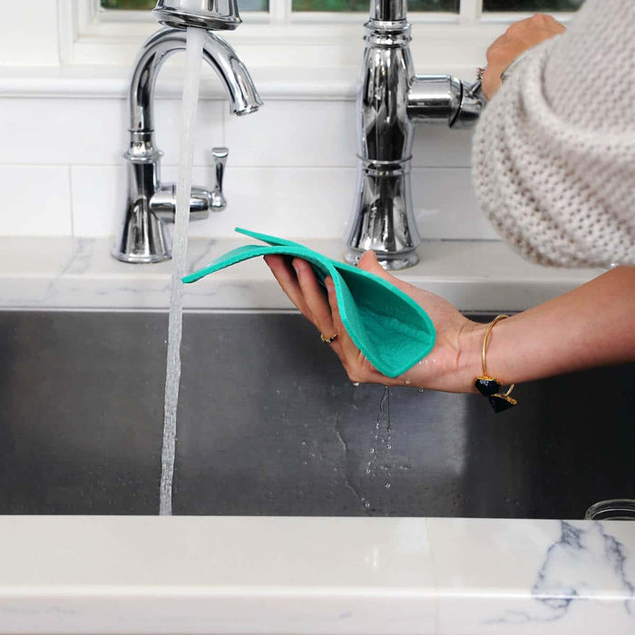 A person's hands holding a folded teal reusable cellulose cloth in a kitchen sink with running water and chrome faucets.