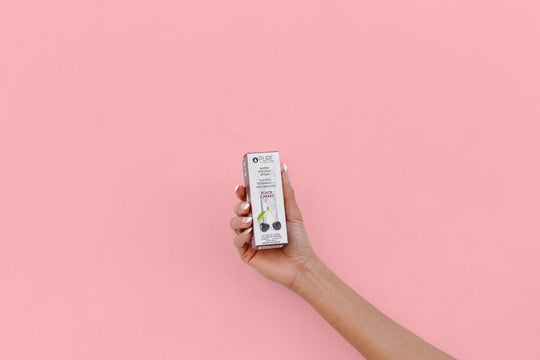 Hand holding a box of Electrolytes + Vitamin C Black Cherry - | Pure Inventions against a solid pink background, highlighting a wellness supplement product.