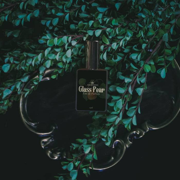 Perfume bottle labeled 'Glass Pear' on a reflective surface with green foliage in the background