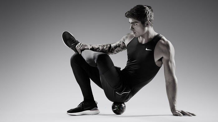 Athletic man using a Hypersphere Go | Hyperice massage ball in black to perform muscle recovery and body care on his leg while wearing workout attire.