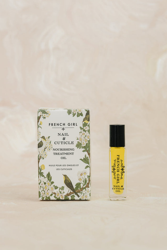 Small glass bottle of Nail & Cuticle Oil | French Girl with black cap next to floral and bird patterned box on a soft beige background, showcasing skincare product.