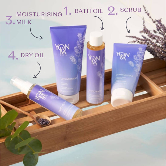 Set of four lavender-scented skincare products on a wooden bath tray including Phyto Bain - Invigorating, Relaxing Aromatic Bath I Yon-Ka Paris bath oil, scrub, moisturizing milk, and dry oil with natural herbs in the background.