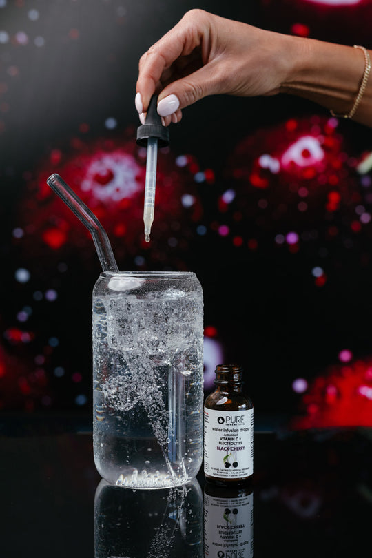 Hand holding dropper releasing liquid into a glass of sparkling water with a clear straw beside a bottle of Electrolytes + Vitamin C Black Cherry - | Pure Inventions against a dark cherry-themed background.