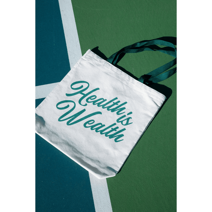 White fabric tote bag with dark green text "Health is Wealth" and matching dark green handles, laid on a green and blue surface, Health Is Wealth Tote | Lucky Owl.