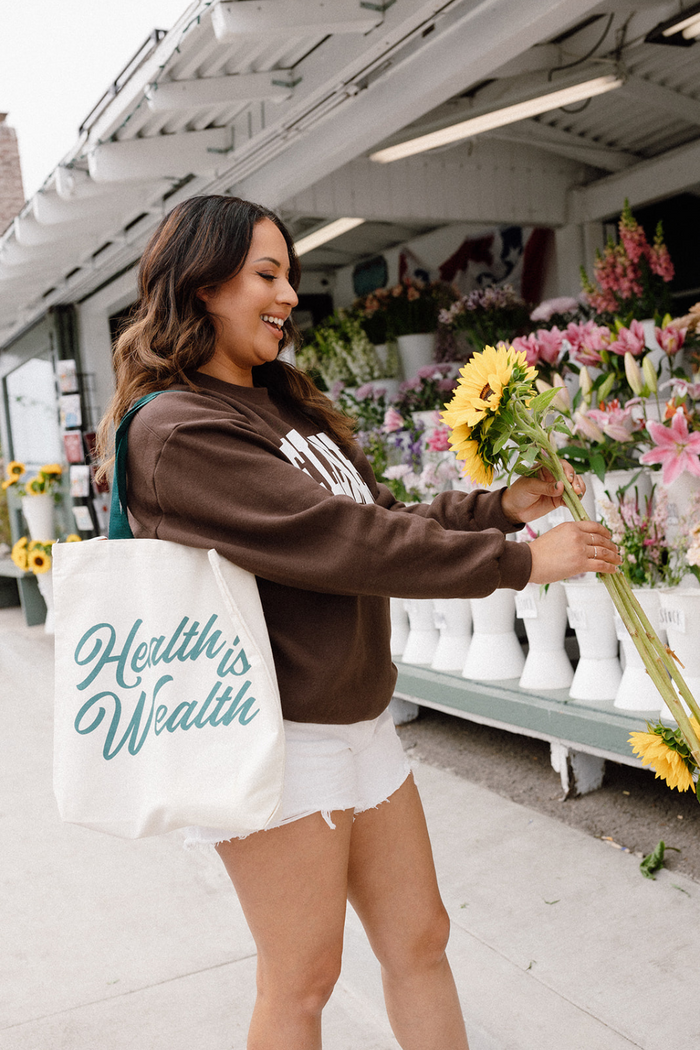 A smiling woman holding sunflowers and wearing a casual brown sweatshirt and white shorts, carrying a canvas tote bag with teal script that reads Health Is Wealth Tote | Lucky Owl at a flower stand.