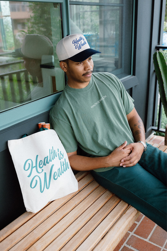 Man wearing green shirt and a baseball cap sitting on wooden bench next to a canvas tote bag with "Health Is Wealth Tote | Lucky Owl" script in teal lettering beside him.