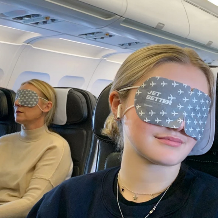 Two women sitting on an airplane wearing gray Jet Setter Warming Eye Mask - Single | Popmask with white airplane patterns, promoting relaxation and wellness during travel.
