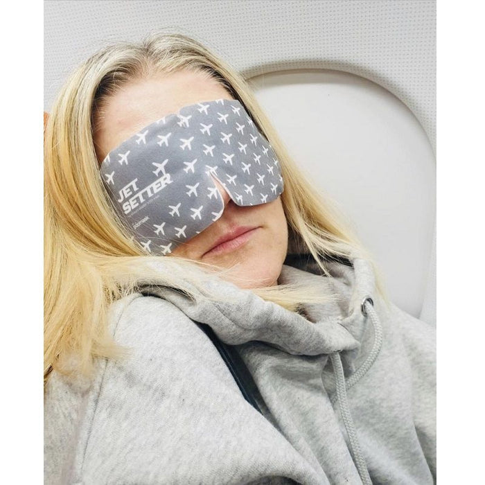 A person wearing a gray Jet Setter Warming Eye Mask - Single | Popmask with white airplane patterns, resting comfortably on an airplane seat while dressed in a light gray hoodie.