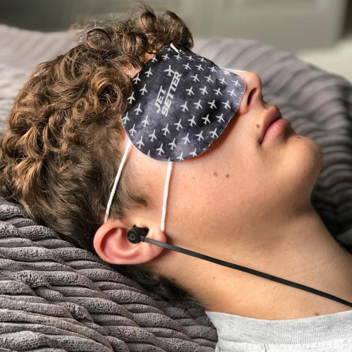 Man lying down wearing a Jet Setter Warming Eye Mask - Single | Popmask with airplane pattern and "Jet Setter" text while relaxing with earbuds.