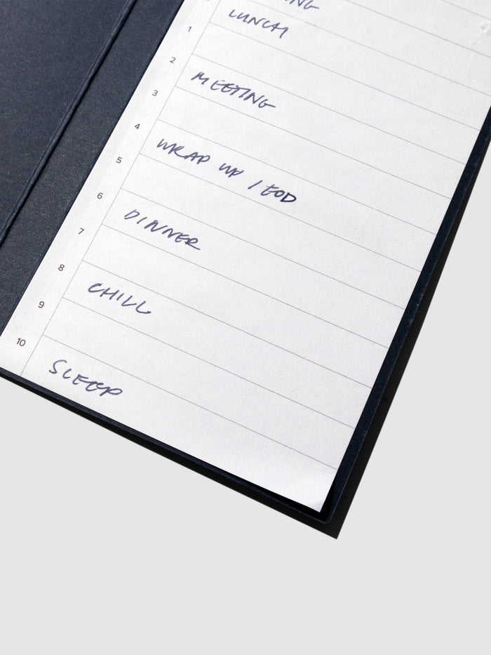 Closeup of sticky note displayed on its backing on a neutral background. It is filled out with sample text showcasing a schedule with a meeting, end of day wrap up, dinner, relaxation time, and sleep.
