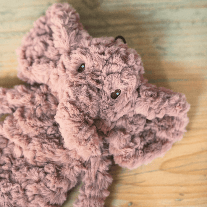 Close up of dusky pink elephant shaped dog toy with long arms and legs, stitched detail eyes on a wooden floor.