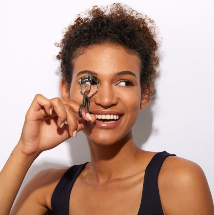 Woman with curly hair smiling while using the Pro Master Lash Curler | Tweezerman to curl her eyelashes against a white background