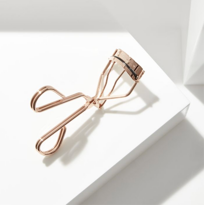 Procurl Lash Curler | Tweezerman in a metallic rose gold finish placed on a white geometric surface with soft natural lighting highlighting the sleek design.