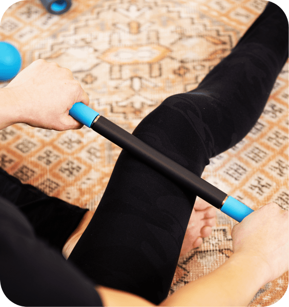 Person using a Rod Mini | RAD massage tool with blue handles on their leg while sitting on a patterned rug, promoting muscle recovery and wellness.