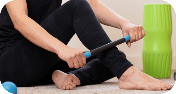 Person using the Rod Mini | RAD massage tool to relieve foot and leg muscle tension while sitting on the floor near a green foam roller in a wellness setting.