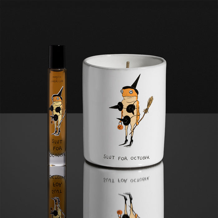 A white ceramic candle jar and a small clear glass roller bottle, both printed with a whimsical Slut for October, on a dark reflective surface against a black background.