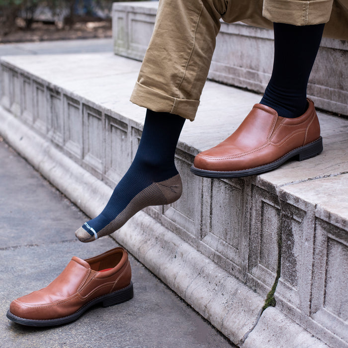 Knee-high compression sock in navy worn with brown slip-on shoes, resting on a stone step.