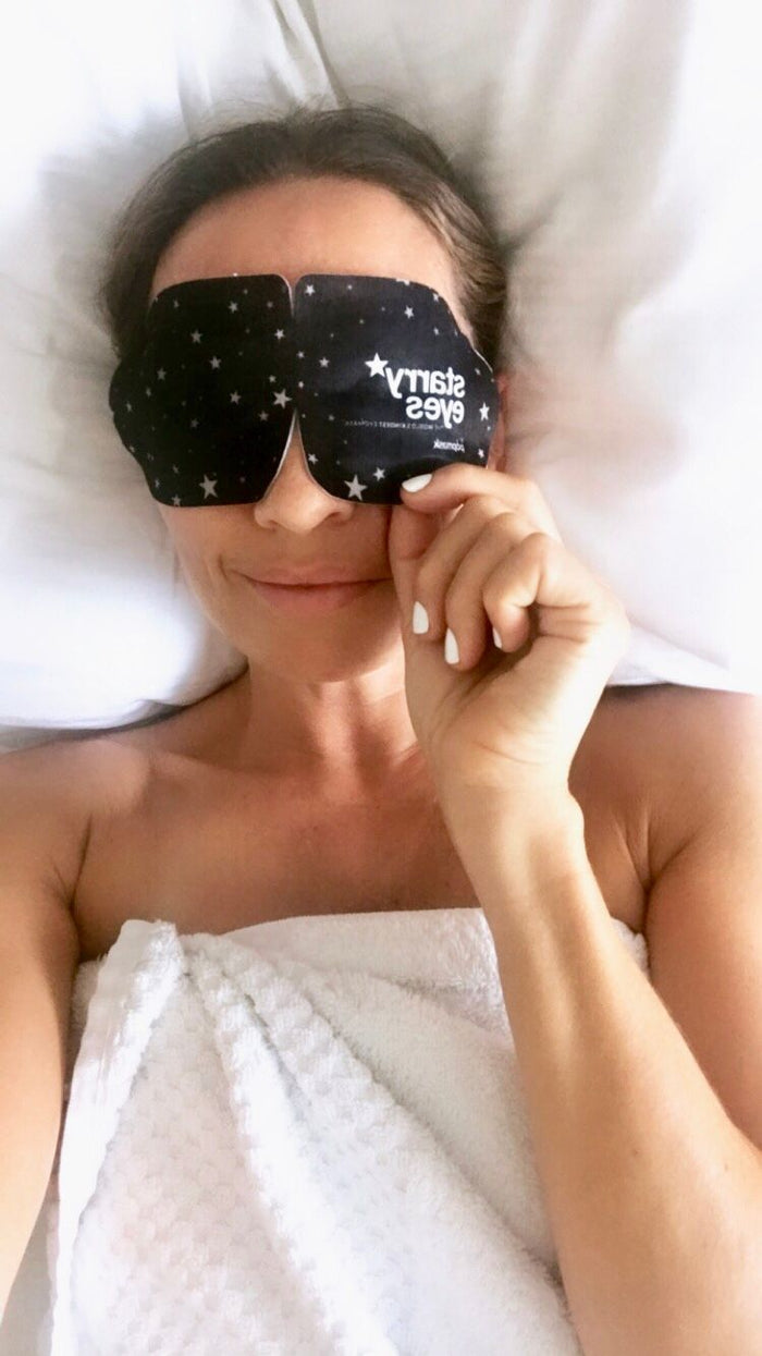 Woman lying on a bed with a white towel wrapped around her wearing a black Starry Eyes Warming Eye Mask - Single | Popmask decorated with small stars, promoting relaxation and skincare wellness.