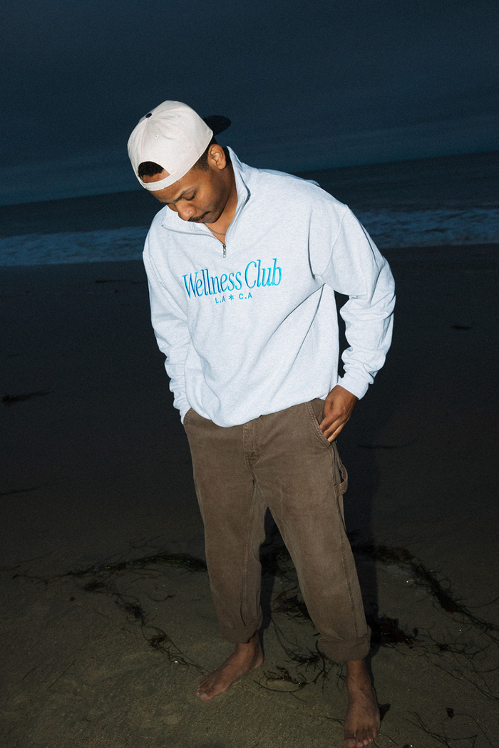 Man standing barefoot on the beach at dusk wearing a light gray quarter zip pullover with blue "Wellness Club L.A. C.A" text and brown pants, styled with a white baseball cap, showcasing the Wellness Club Quarter Zip | Lucky Owl.